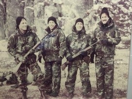 group of men standing in camouflage uniforms in cold weather two men are holding rifles and there is snow laying on the ground and on the branches of the trees