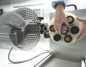 Les mains d’un technicien placent des récipients à l’intérieur d’un dispositif relié à un ventilateur entouré d’une grille métallique.