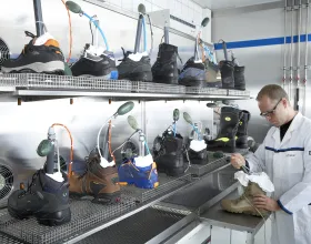 Un homme en blouse blanche fait un test sur une chaussure. Autour de lui, plusieurs chaussures en test sur des étagères métalliques.